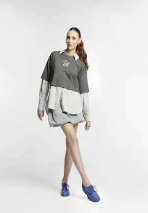 Top gris de manga corta con diseño bordado superpuesto a una camisa de botones gris claro y pantalones cortos grises, combinado con zapatos azul con cordones.