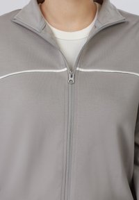 Gros plan d'une personne portant une veste zippée gris clair avec une ligne horizontale blanche et un t-shirt côtelé blanc en dessous.