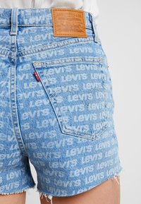 Lähikuva vaaleansinisistä farkkushorteista, joissa on toistuva "Levi's" -tekstikuvio, takatasku, punainen Levi's -merkki ja ruskea nahkainen logopatch.