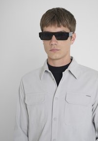 Gafas de sol negras con un marco rectangular, llevadas por una persona con una camisa de pana gris claro con dos bolsillos en el pecho y una camiseta de cuello redondo debajo.