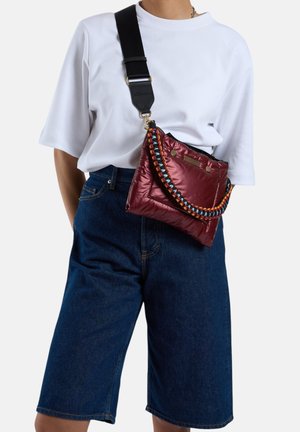 Sac matelassé rouge avec une sangle tressée multicolore et une bandoulière noire réglable. Porté en bandoulière avec un t-shirt blanc ample et un short en jean.