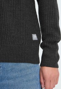 Sort strikket sweater med ribbestruktur, der har et lille mærketik nær sømmen og et tætsiddende manchet-design.