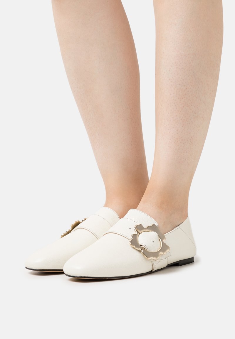 Weiße Leder-Slipper haben eine spitze Zehenpartie, einen Riemen über dem Mittelfuß und eine dekorative goldene Hardware-Akzentuierung. Flache Sohle.
