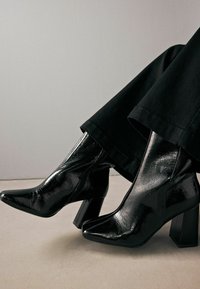 Stivaletti alla caviglia in pelle verniciata nera con punta affusolata e tacco a blocco spesso, caratterizzati da una superficie testurizzata e un design elegante.