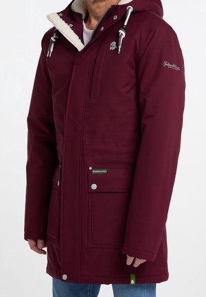 Mężczyzna w bordowej zimowej kurtce z kapturem, z podszyciem z polaru, przednimi kieszeniami oraz naszywkami z logo na rękawie i kieszeni.