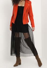 Veste de moto en suède rouge sur une robe noire avec un superposé en dentelle transparente, associée à des bottes en suède marron jusqu'au genou.