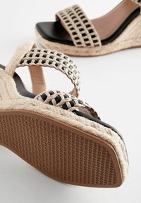 Sandales à talon compensé avec des sangles en rotin noires sur une base beige, des détails en jute tressé et une semelle extérieure en caoutchouc brun texturé.