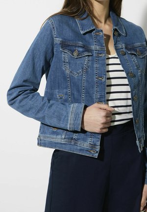 Femme portant une veste en jean bleue par-dessus une chemise blanche et noire à rayures, associée à un pantalon bleu marine foncé, debout devant un fond uni.