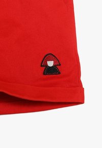 Czerwone szorty z materiału mają podwinięty brzeg i czarne haftowane logo grzyba w dolnym rogu, ukazując gładką fakturę i intensywny kolor.