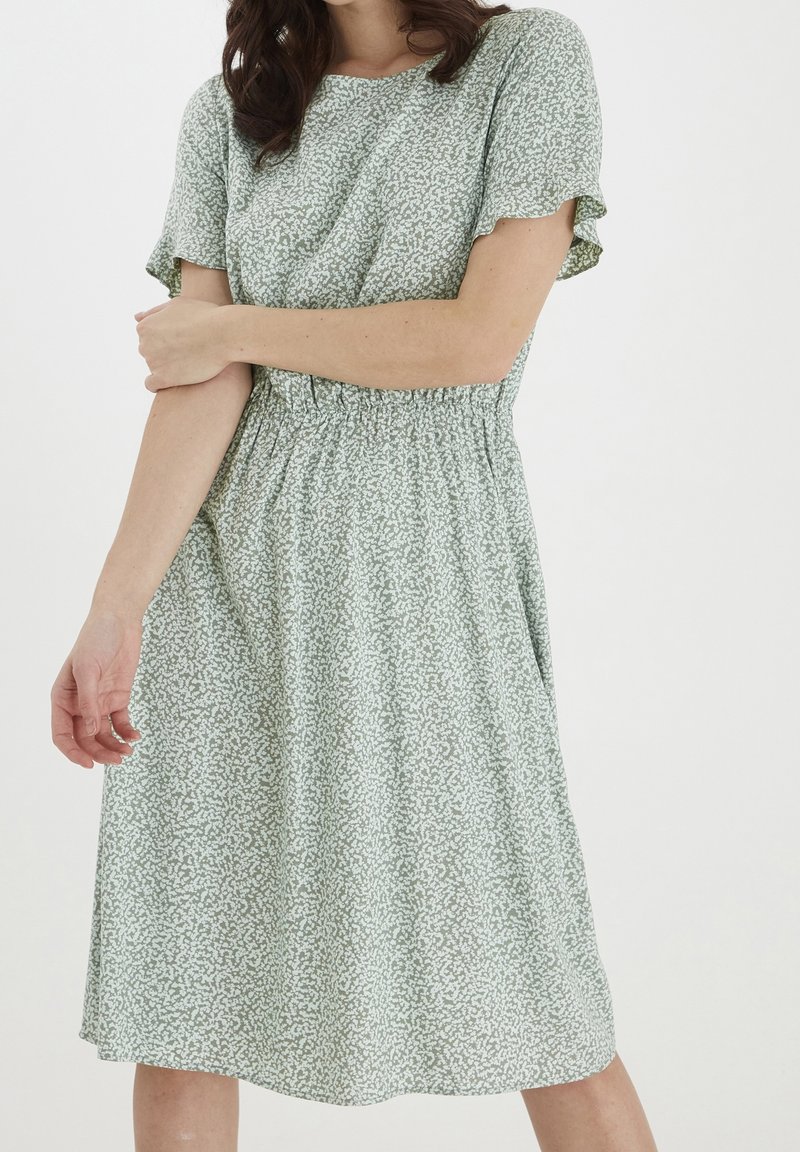 Robe verte à motifs avec des manches courtes, taille élastique, tissu fluide et coupe mi-longue arborant un subtil design floral blanc.