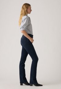 Dark blue flared jeans with a high waist, made from denim, paired with a striped button-up shirt. Model stands in profile.