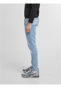 Hele sinised kitsad teksad, tehtud denimist, kus on kulunud detailid ja klassikaline viie taskuga disain, kombineeritud hallide spordikingadega.