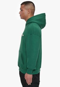 Grøn hættetrøje lavet af blødt stof, med en kængurulomme, ribbede håndled og en hætte med snor. Minimalistisk design uden synlige mønstre.