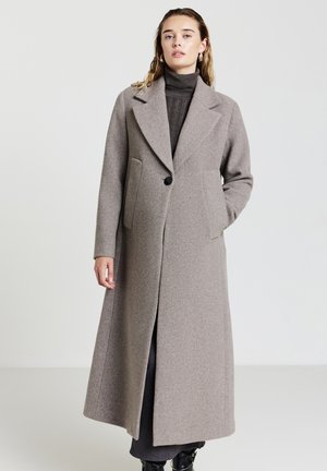 Donna che indossa un lungo cappotto di lana taupe sopra un maglione a collo alto scuro, in piedi con una mano in tasca su uno sfondo neutro.