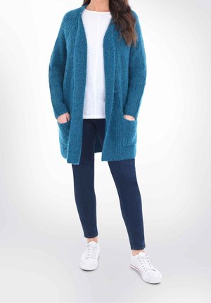 Mujer que lleva un cárdigan abierto de color verde azulado sobre una camisa blanca, vaqueros azul oscuro y zapatillas blancas, de pie con las manos en los bolsillos del cárdigan.