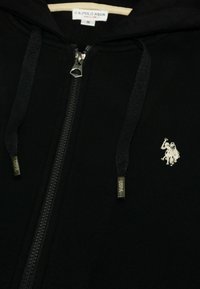 Fekete cipzáras kapucnis pulóver, puha anyagból készült, bordázott textúrával. Ezüst cipzárral és egy kis hímzett logóval rendelkezik a bal mellen.