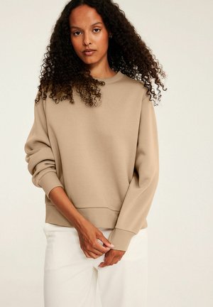 Kvinde med krøllet hår iført beige sweater og hvide bukser står mod en ensfarvet lys baggrund, hænderne blidt foldet foran.