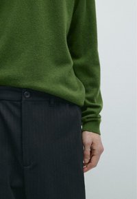 Maglione verde in maglia con polsini a coste abbinato a pantaloni scuri a righe sottili. La mano si posa sulla vita dei pantaloni, mostrando i dettagli dei bottoni.