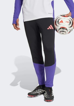 Person in schwarzen und violetten Adidas Sporthosen hält einen weißen Adidas Tiro Fußball und trägt schwarze Stollenschuhe mit roten Nocken.