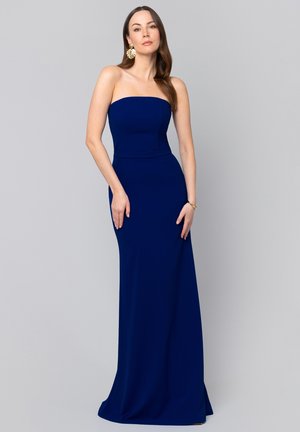 Kobieta z długimi brązowymi włosami, ubrana w granatową, długą suknię bez ramiączek oraz złote kolczyki w kształcie liści, stojąca na tle jednolitego, szarego tła.