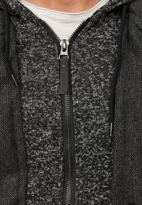 Zwarte en grijze zip-up hoodie met een textuur gebreid patroon, voorzien van een frontale rits, trekkoordkap en een subtiele merketiket.
