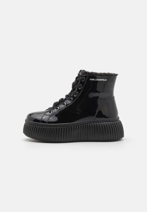 Black patent leather ankle boots with a textured, ridged sole, featuring a fur collar and lace-up design. Logo on the side is visible.