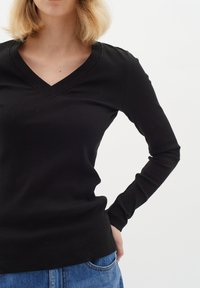 Top nero a maniche lunghe con scollatura a V, design aderente, texture liscia e dettagli di cucitura sottili. Indossato con jeans in denim.