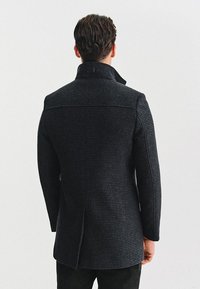 Czarnoszary płaszcz wełniany z teksturowanym wzorem, z wysokim kołnierzem i prostym designem z tyłu. Posiada wentylację z tyłu dla większej swobody ruchów.