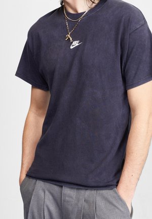 Personne portant un t-shirt Nike bleu foncé et un pantalon gris, les mains dans les poches, accessoirisée avec des colliers chaînes dorés superposés.