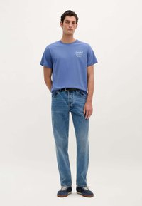 Tricou din bumbac albastru cu mâneci scurte, cu un imprimeu logo pe piept, combinat cu blugi drepti de culoare albastru deschis și adidași negri.