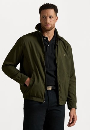 Olive green jacket with a zip front, stand collar, and elastic hem. Features side pockets and a small logo on the chest. Worn over a black polo.
