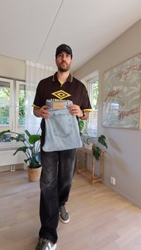 Jeans de mezclilla de color azul claro con una etiqueta, sostenidos por una persona en una camiseta polo marrón con acentos amarillos. El fondo incluye plantas y una obra de arte en la pared.