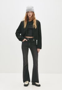 Ung kvinna iklädd en grå mössa, svart kort jacka, svart kort topp, mörka utsvängda jeans och svart-vita loafers som står framför en neutral bakgrund.