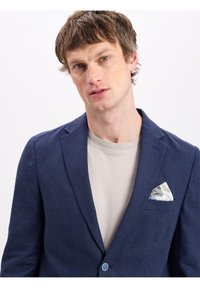 Marineblauw linnen blazer met één knoop, vergezeld van een lichtbeige t-shirt. Bevat een gebloemde witte pochet en gestructureerde stof.