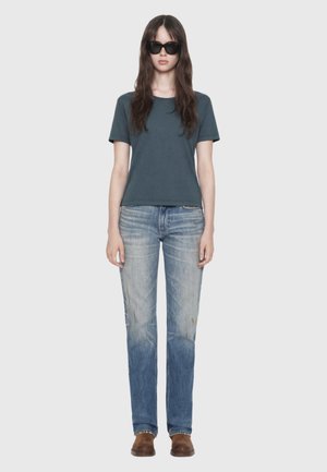 Woman with long brown hair wearing black sunglasses, a dark gray short sleeve t-shirt, faded blue jeans, and brown shoes, standing still.
