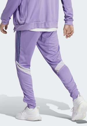 Personne portant un pantalon de survêtement violet avec des rayures blanches et bleu foncé et des baskets blanches, debout avec une jambe tendue vers l’arrière.