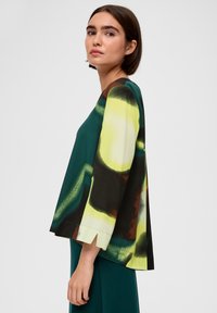 Felgekleurde top met groene en gele abstracte ontwerpen, voorzien van vleermuismouwen en een recht gesneden silhouet. Gelaagd stof.