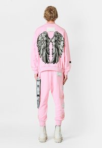 Sudadera rosa con un gran bordado de alas plateadas en la parte posterior, combinada con joggers rosa desgastados a juego y botas blancas.