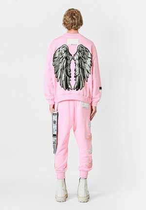 Sudadera rosa con un gran bordado de alas plateadas en la parte posterior, combinada con joggers rosa desgastados a juego y botas blancas.