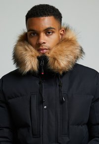 Schwarze Steppjacke mit einem großen, abnehmbaren Kragen aus tanfarbigem Fell. Verfügt über Reiß- und Druckknopfverschlüsse, verstellbare Zugbänder und Fronttaschen.
