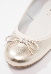 Gouden metallic ballerina met een afgeronde neus, witte elastische rand en een kleine strikdetail aan de voorkant. Gladde textuur met glanzende afwerking.