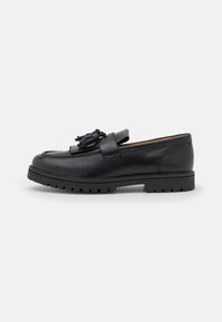 Des mocassins en cuir noir présentent une semelle en caoutchouc à relief, des détails en couture et une frange décorative sur le devant pour une texture ajoutée.