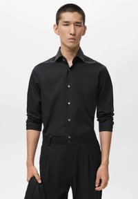 Camisa negra de manga larga con cuello puntiagudo, con una parte delantera abotonada y un brillo sutil. Llevada con pantalones negros a juego.