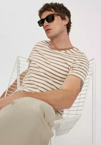 Pasiasta koszulka w odcieniach beżu i brązu, okrągły dekolt, krótkie rękawy. Model nosi czarne okulary przeciwsłoneczne, siedząc na białym drucianym krześle.