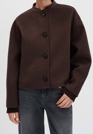 Brown buttoned wool jacket with long sleeves worn with dark gray jeans, hand resting on hip, neutral background.