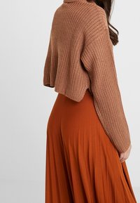 Pull court tricoté marron avec manches larges, associé à une longue jupe plissée orange fluide. Texture douce, palette de couleurs chaleureuse.