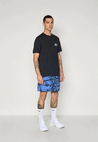 Marineblaues T-Shirt mit einem kleinen türkisfarbenen Logo, kombiniert mit blauen Batikshorts, weißen Socken und weißen Sneakern. Modell steht aufrecht.