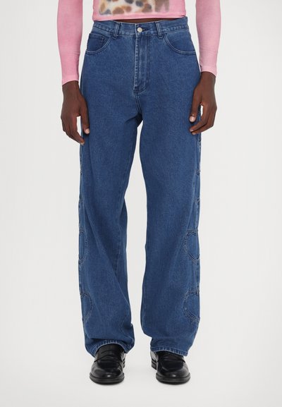 Light blue denim jeans with a loose fit, featuring side button accents and curved cut-out details along the outer seam.