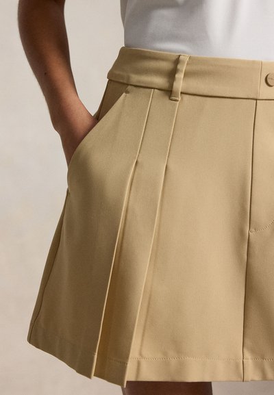 Φούστα ανοιχτού καφέ με πιέτες και ψηλή μέση, θηλιές για ζώνη, πλαϊνή τσέπη, φορεμένη με λευκό τοπ, χέρι που στηρίζεται στην τσέπη.