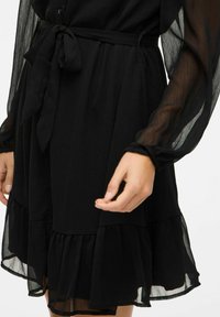 Abito nero con maniche lunghe trasparenti, vita con lacci e orlo a strati con balze. Il tessuto ha un aspetto leggero e testurizzato.